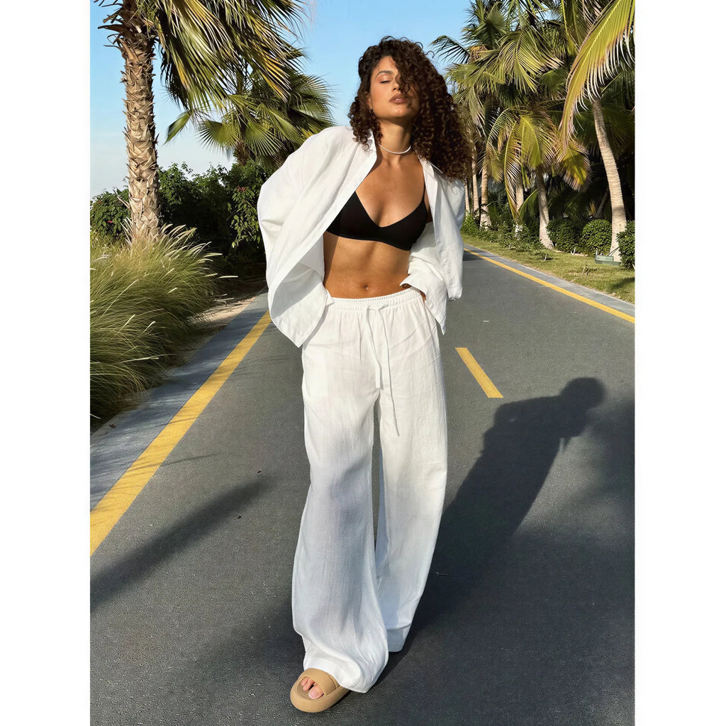 Woman wearing a white long sleeve fabric shirt and matching drawstring trousers over a black top on a paved road.