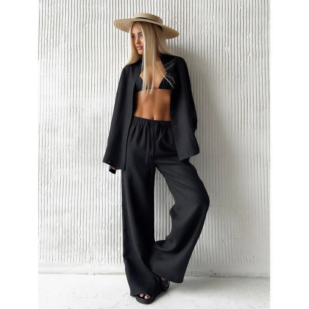 Woman wearing a black long sleeve lapel shirt and matching drawstring trousers with a straw hat against a white textured w...