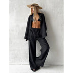 Woman wearing a black long sleeve lapel shirt and matching drawstring trousers with a straw hat against a white textured w...