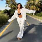 Woman wearing a white long sleeve button-down shirt and matching trousers over a black top on a paved road with palm trees.